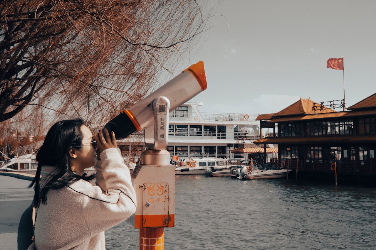 portfolio-07 Asian woman looks through telescope by river with distant landmarks and boats.