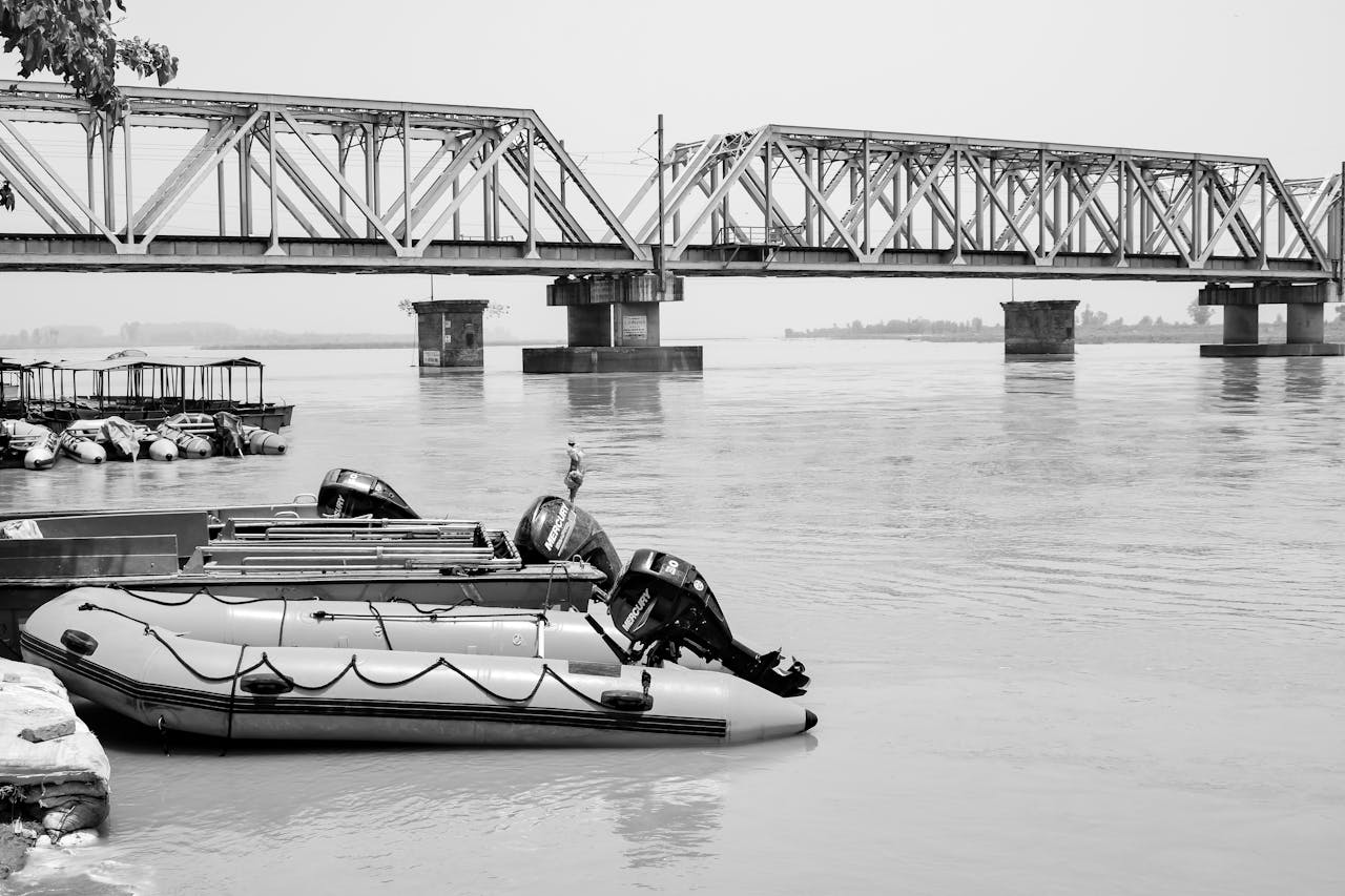 portfolio-05 A black and white image showcasing boats docked by a metal bridge over a calm river.
